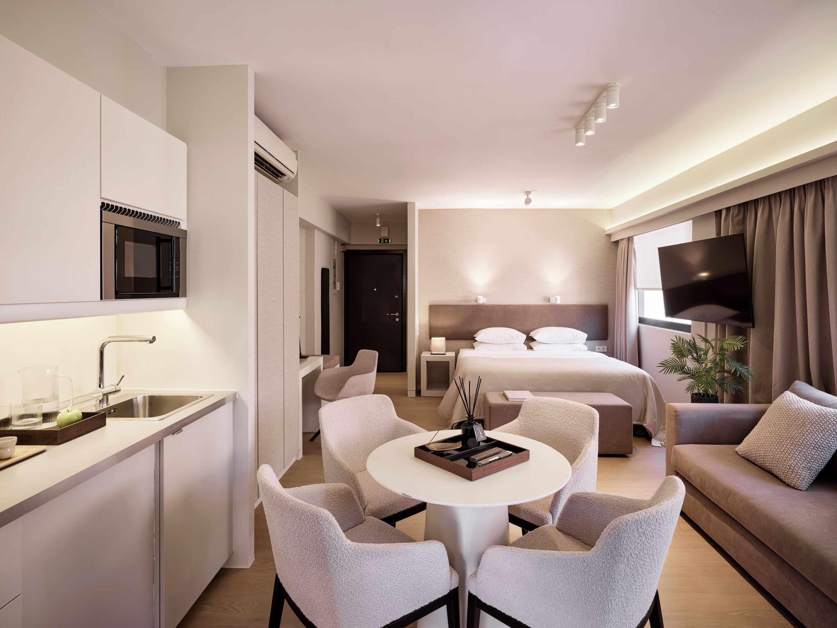 Una habitación de hotel moderna con kitchenette, cama con televisor, zona de estar y un amplio ventanal.