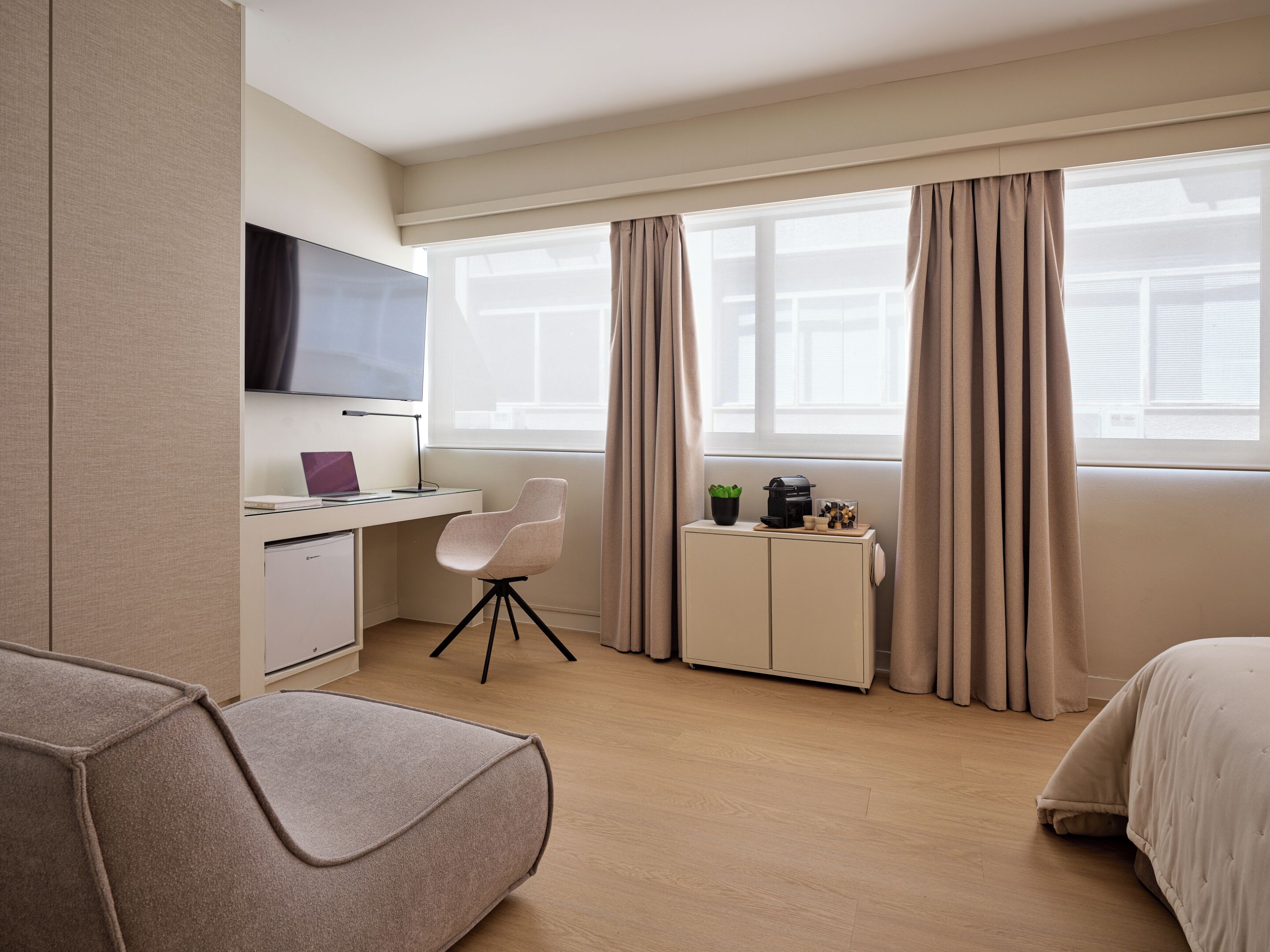 A hotel room with a bed, a desk with a computer, a TV, and a window with curtains.