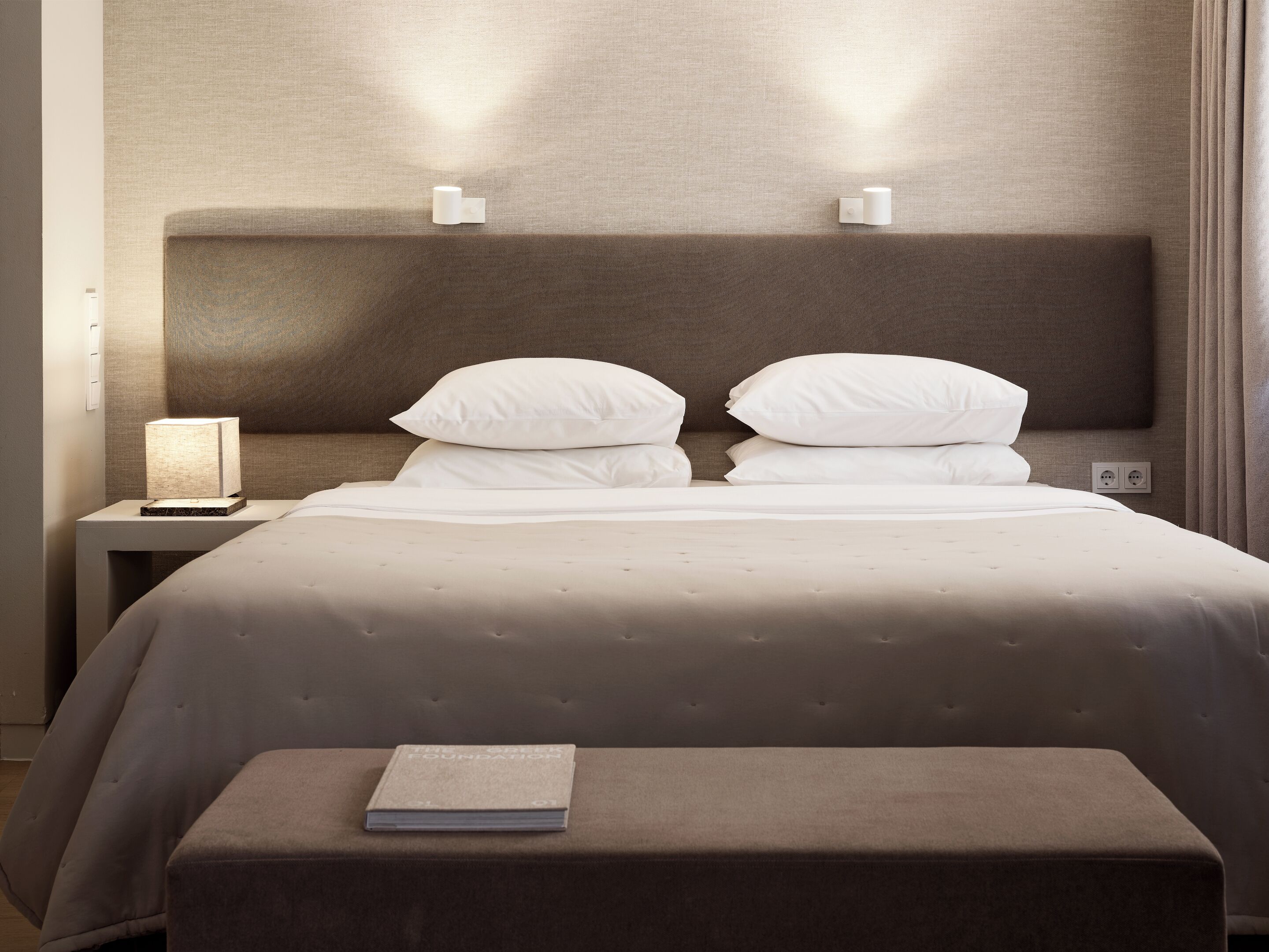 A neatly made bed with a headboard, a bedside lamp, and a book on an ottoman.