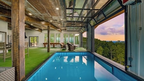 A modern indoor swimming pool with a glass ceiling, wooden beams, and a view of the forest. - Skyline Sanctuary by Avantstay Indoor Pool, Game Room, Firepit, Mountain Views, Luxe Design (Sevierville)