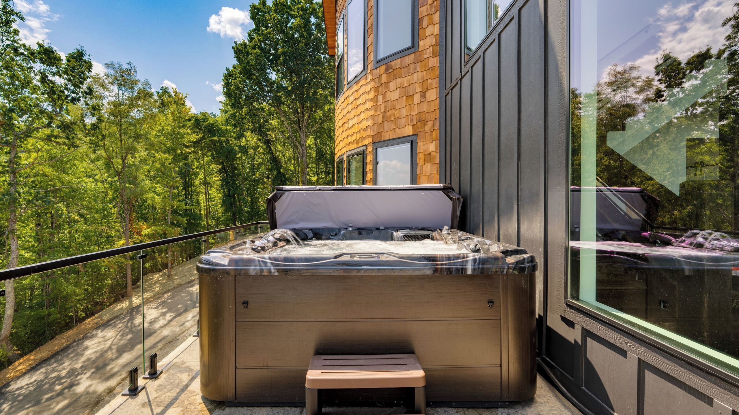 A hot tub with a bench, a glass railing, and a view of trees and a building.