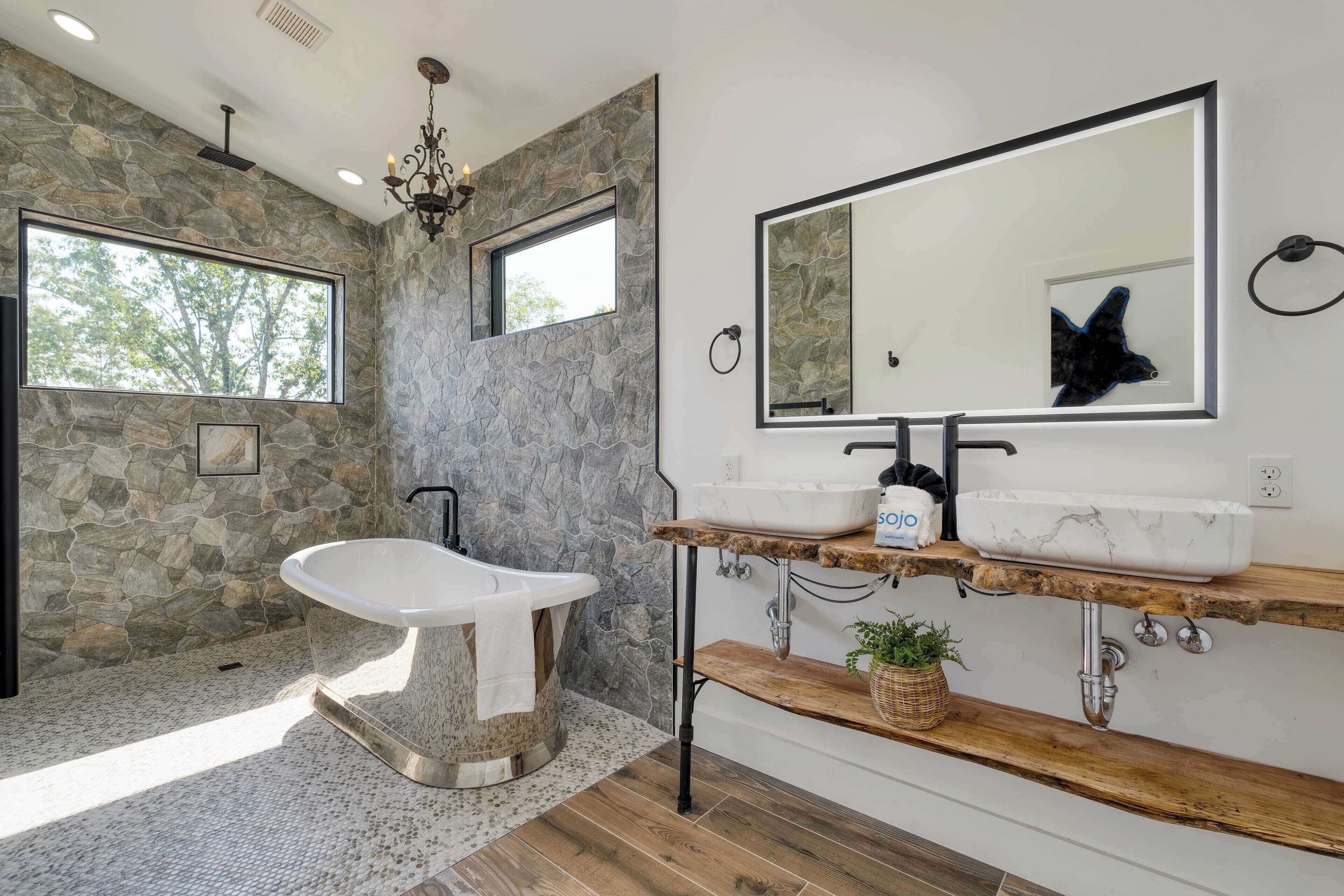 A bathroom with a freestanding bathtub, two sinks on a wooden shelf, a large mirror, and a stone wall.