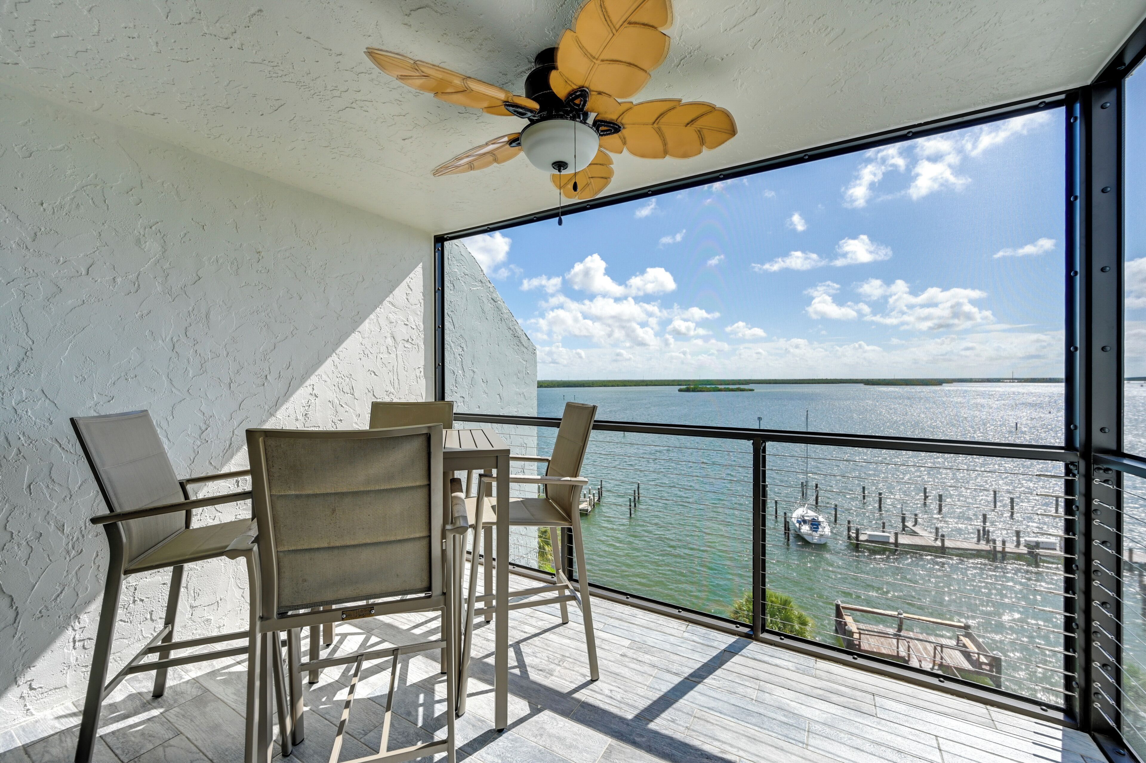 A balcony with a table and chairs, a ceiling fan, and a view of a body of water with boats and docks.