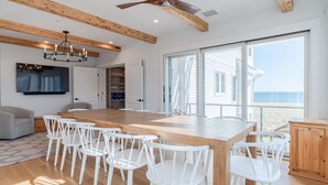 Un comedor amplio con una gran mesa de madera y sillas blancas, un candelabro y vistas al océano a través de grandes ventanales.