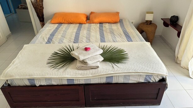 A bed with striped bedding, two orange pillows, a folded towel, and a decorative palm frond. - Esmeralda Djerba Casino (Mezraya)