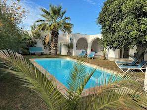 Une piscine entourée de palmiers et de chaises longues, avec un bâtiment aux arches apparentes en arrière-plan.