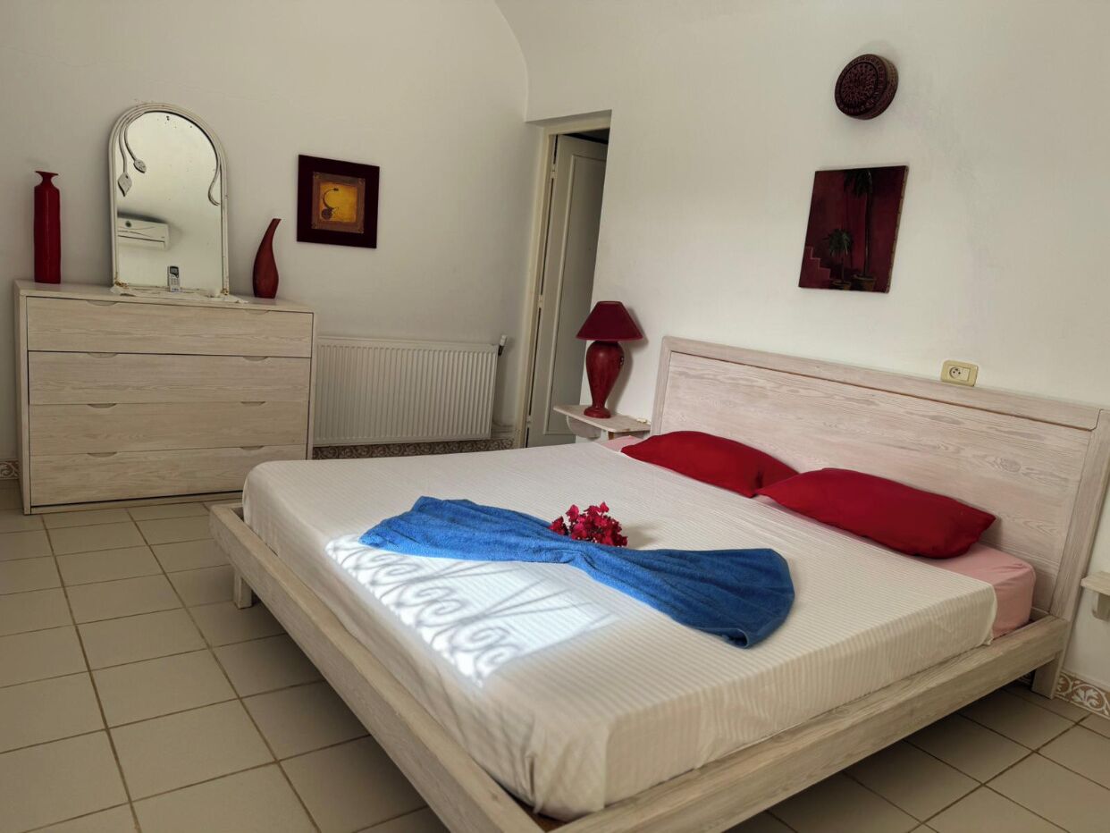 Een net opgemaakt bed met een blauwe handdoek en rode kussens, een houten dressoir met een spiegel, en een rode vaas op het dressoir.