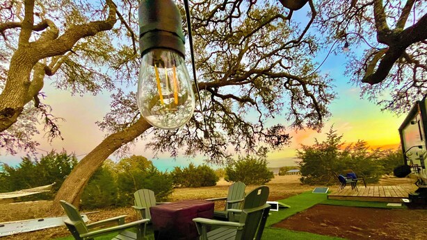 A patio with green chairs and a table, a hanging light fixture, and a view of the sunset. - Ramblin' Rose: a sweet Glamper for 2 under the stars in Dripping Springs (Dripping Springs)
