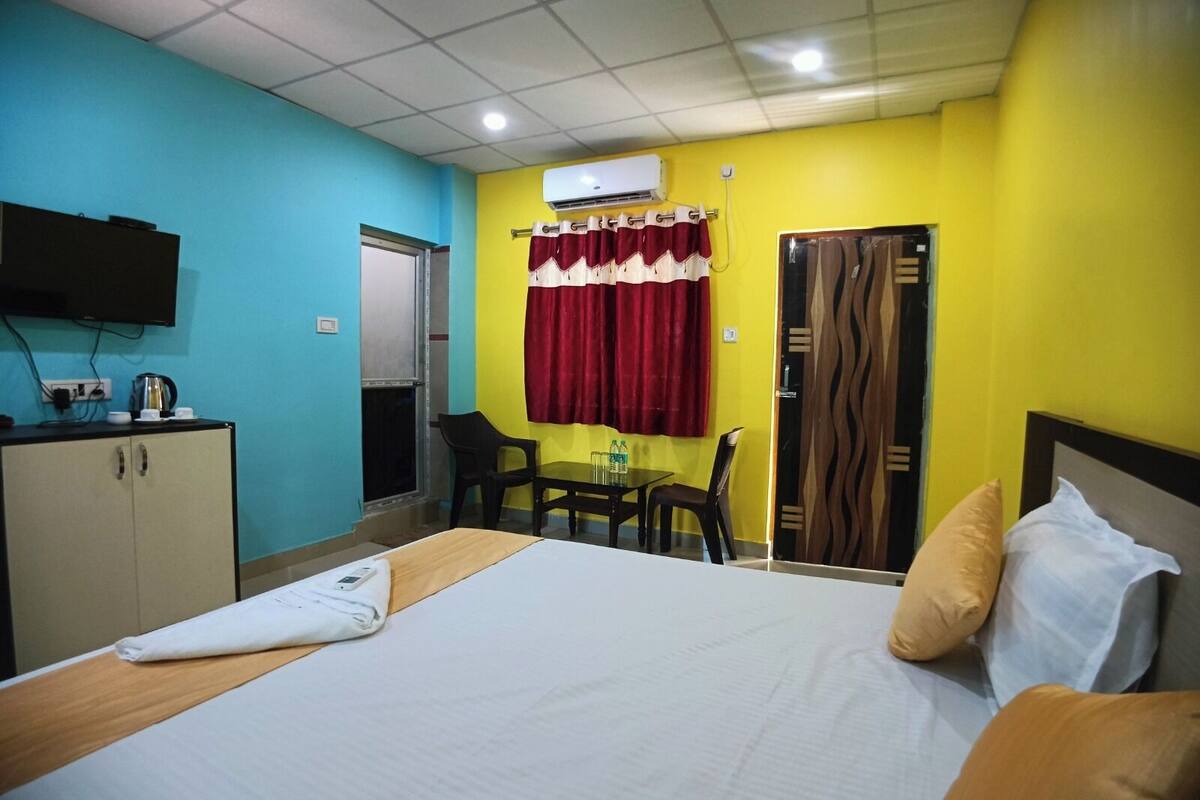A hotel room with a bed, a table with chairs, a TV, and an air conditioning unit.
