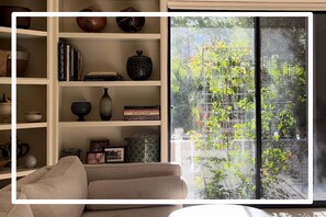Un salón con sofá beige, una estantería decorada y un ventanal con vistas a un jardín.