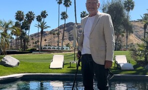 A man standing by a pool with palm trees and a mountain in the background.