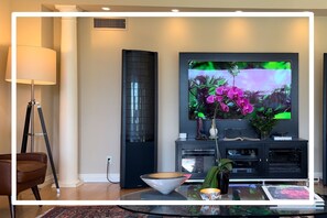 A modern living room with a large flat-screen TV, a floor lamp, a dark-colored entertainment unit, and a glass coffee table.