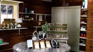 Un intérieur rustique en bois, avec une grande table en bois au centre où sont exposés divers produits, des étagères remplies d’articles et un ventilateur de plafond.