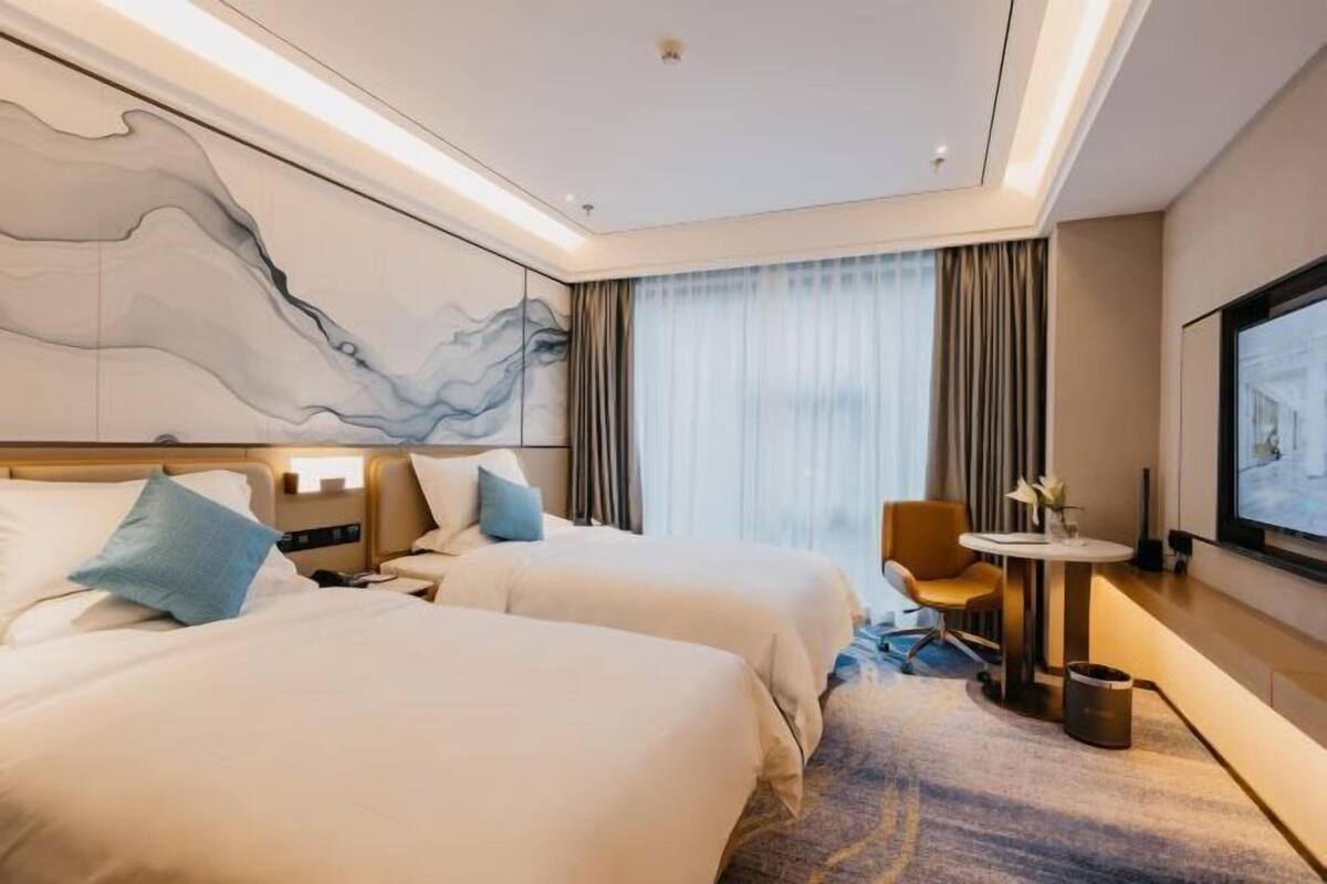 Ein Hotelzimmer mit zwei Betten, einem Schreibtisch, einem Sessel, einem Fernseher und einem großen abstrakten Gemälde an der Wand.