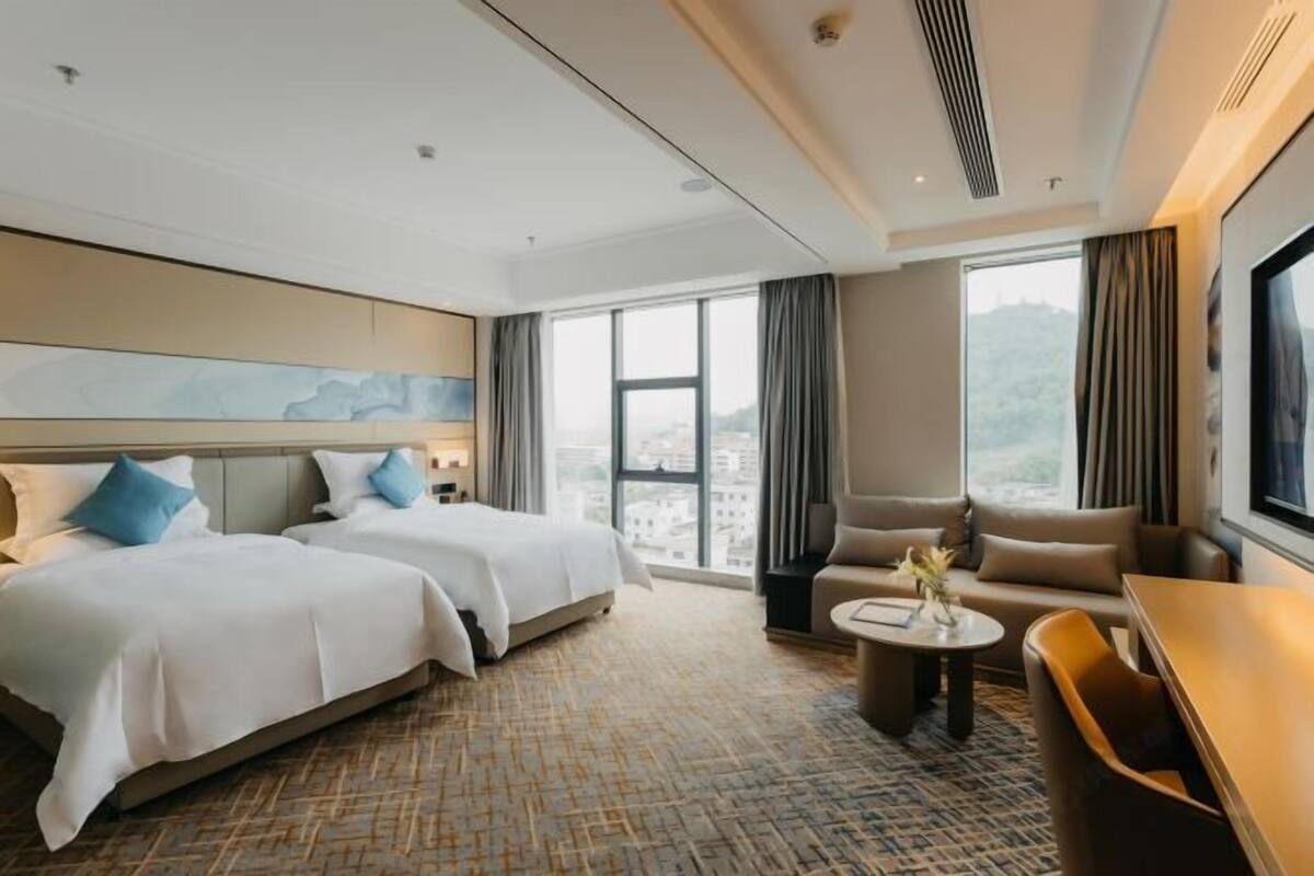 Ein Hotelzimmer mit zwei Betten, einem Sofa, einem Couchtisch und einem großen Fenster mit Blick auf die Stadt.