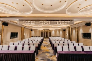 Un salón de conferencias con mesas dispuestas en filas, una araña de cristal y una pantalla de televisión.