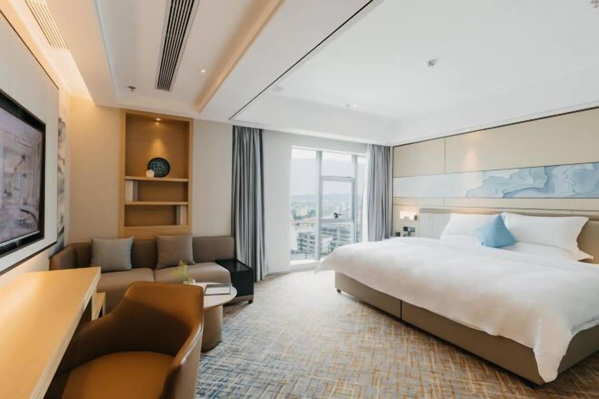 Ein modernes Hotelzimmer mit einem großen Bett, einer Sitzecke, einem Schreibtisch und Blick auf die Stadt durch das Fenster.