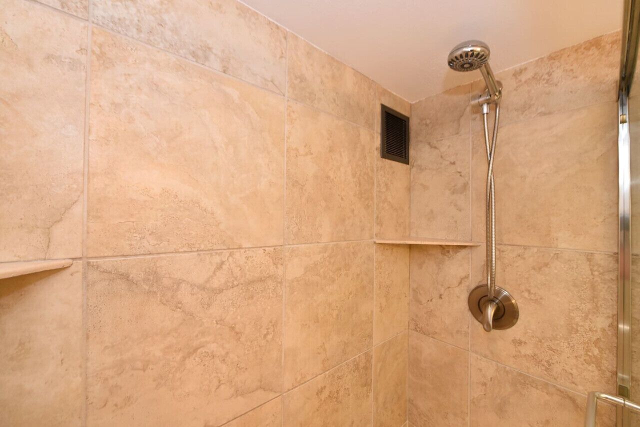 A bathroom with beige marble tiles, a shower with a handheld showerhead, and a small ventilation window.