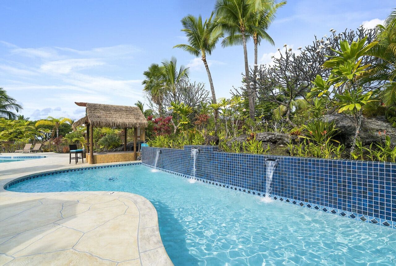 A swimming pool with a blue tiled edge, surrounded by tropical vegetation and a thatched-roof structure.