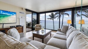 A living room with a large TV, comfortable seating, a coffee table, and a view of the ocean through large windows. - Ocean Front - Gorgeous Views! (Lahaina)