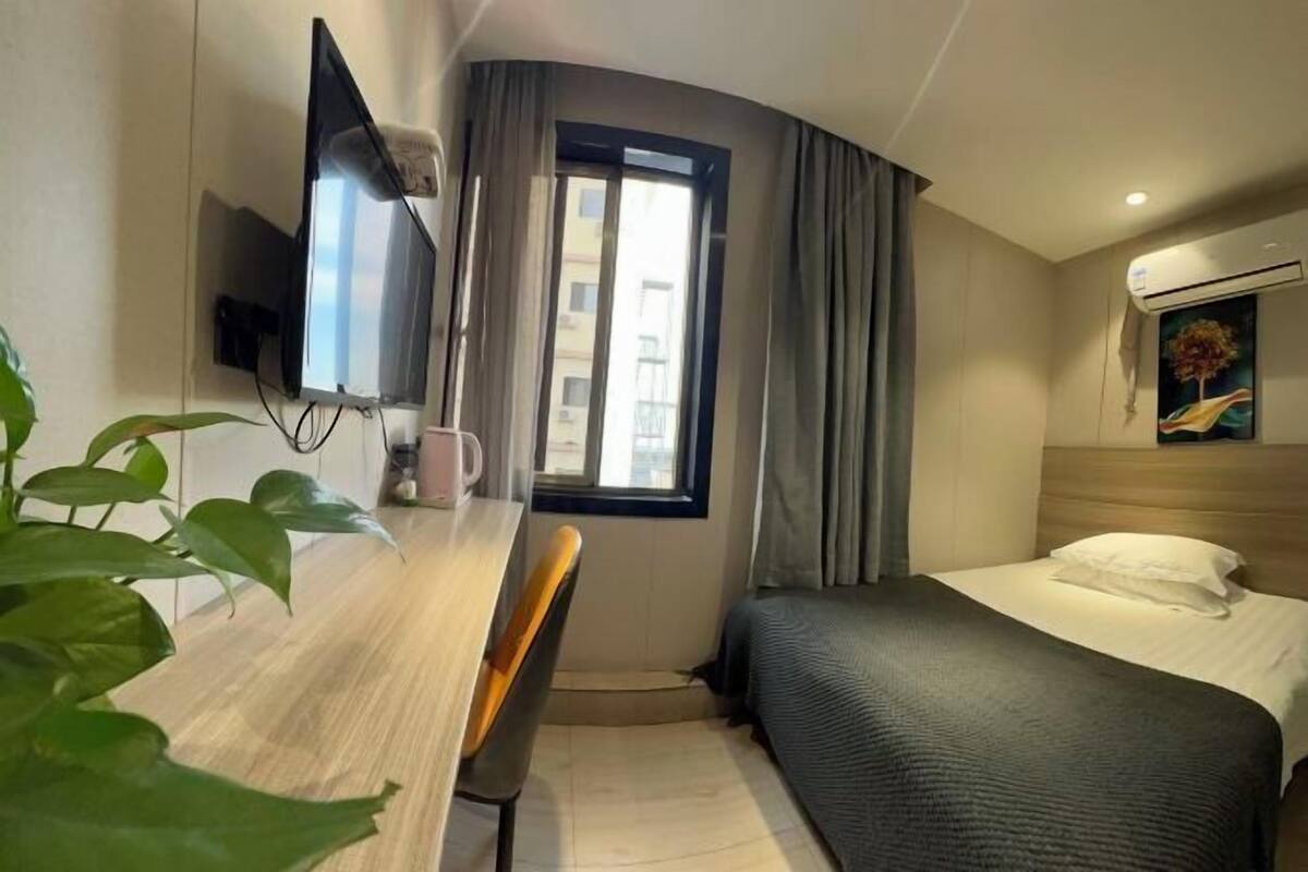 Une chambre d’hôtel comprenant un lit, un bureau avec une chaise, un téléviseur fixé au mur, une fenêtre avec des rideaux et un appareil de climatisation.