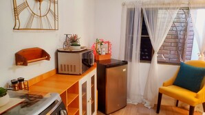 Une cuisine compacte équipée d’une cuisinière, d’un four à micro-ondes et d’un réfrigérateur. On y trouve une étagère en bois ornée d’une petite plante et d’une décoration représentant un vélo. Une chaise jaune est visible près d’une fenêtre avec des rideaux légers.