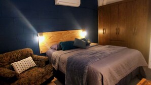 Une chambre à coucher comprenant un lit, une tête de lit en bois, une table de chevet, une lampe, une armoire et un fauteuil à motifs.