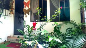 Une maison avec une porte d’entrée, des fenêtres avec des rideaux, un vase rouge et diverses plantes.