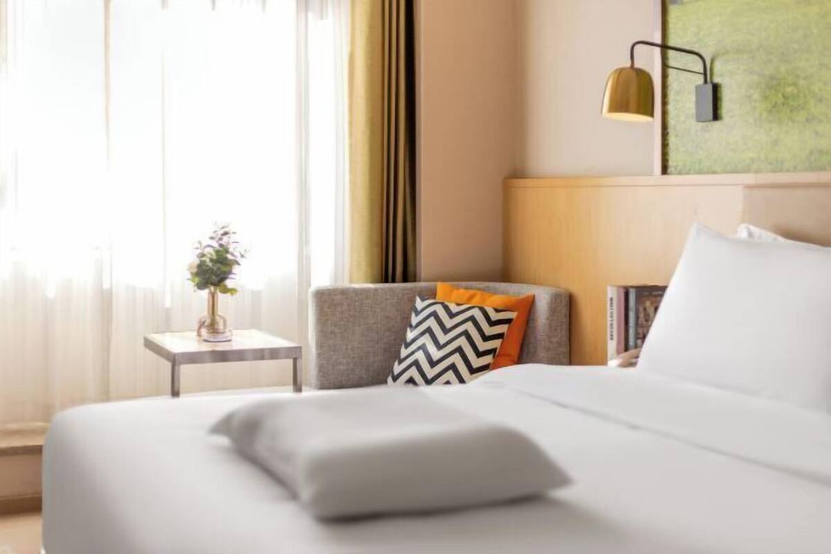 Hotellihuone, jossa on sänky, yöpöytä, jossa on kasvi, lamppu ja seinälle ripustettu taideteos.