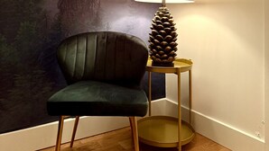 A room with a forest-themed wallpaper, a green armchair, a lamp with a pine cone, and a framed deer picture.