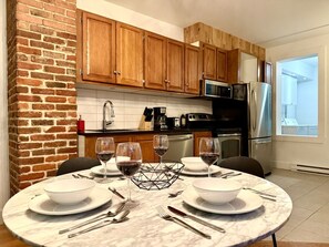A modern kitchen with wooden cabinets, a brick accent wall, and a dining area set for four with wine glasses and plates. - 1277 3rd Avenue · Spacious apartment with parking in Limoilou (Québec)