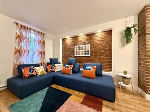 Ein modernes Wohnzimmer mit einer Eckcouch, gemusterten Kissen, einem Teppich, einer Ziegelwand und einem kleinen Beistelltisch mit einer Pflanze.