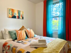Ein Hotelzimmer mit Bett, Kissen, einer gesteppten Decke, einem Fenster mit Vorhängen und einem Bild an der Wand.