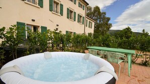 Un bain à remous extérieur, agrémenté d’une table et de chaises, entouré de verdure et adossé à un bâtiment aux volets verts.