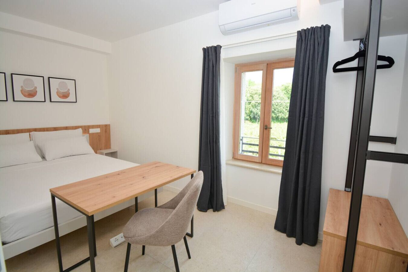 A room with a bed, a wooden table, a chair, a window with curtains, and a wall-mounted air conditioner.