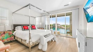 A modern bedroom with a four-poster bed, a large window with a view of a body of water, and a colorful artwork on the nightstand. - Specials Barbie themed, Lakefront, games+pool (Indio)