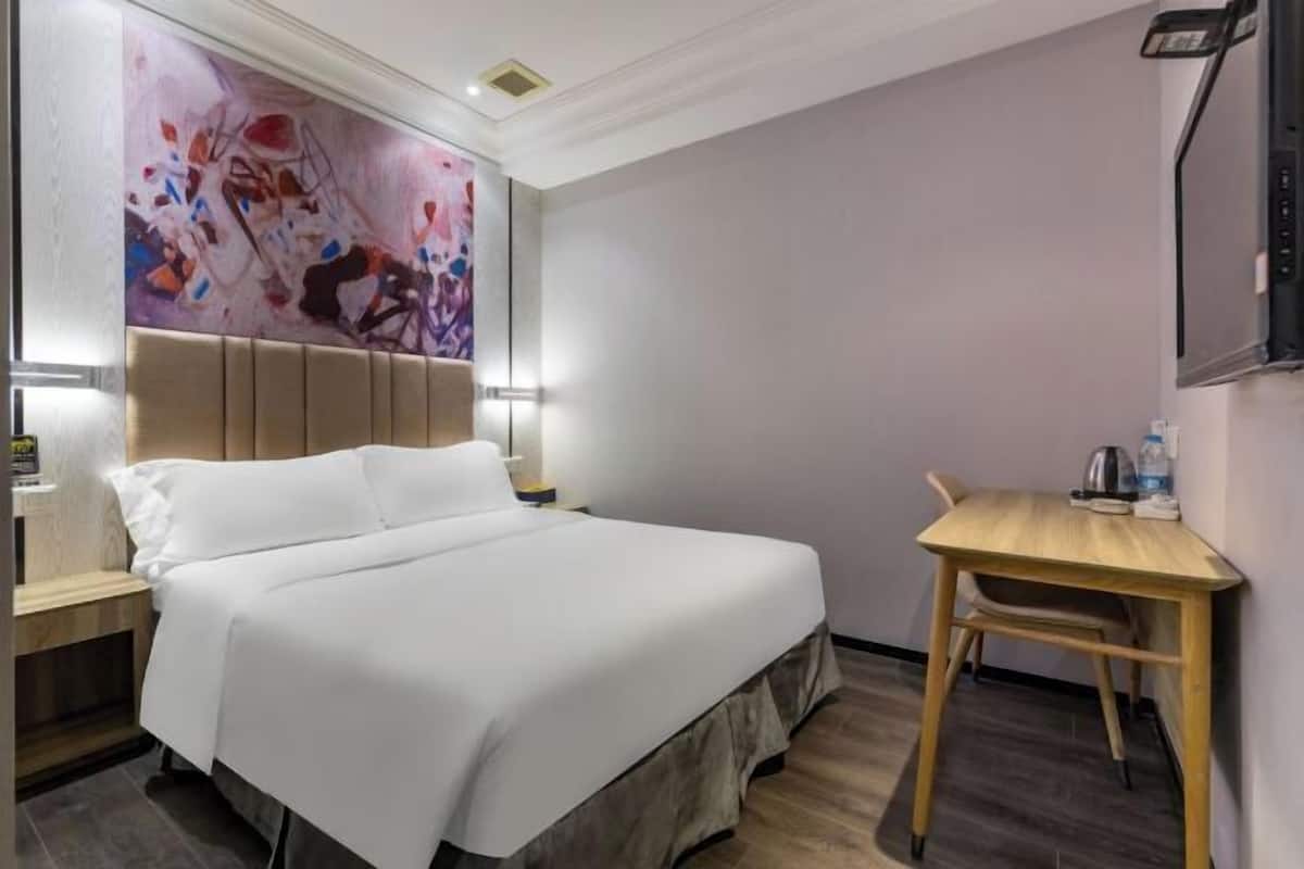 Hotellihuoneessa on sänky, yöpöytä, pieni puupöytä ja suuri, abstrakti maalaus seinällä.