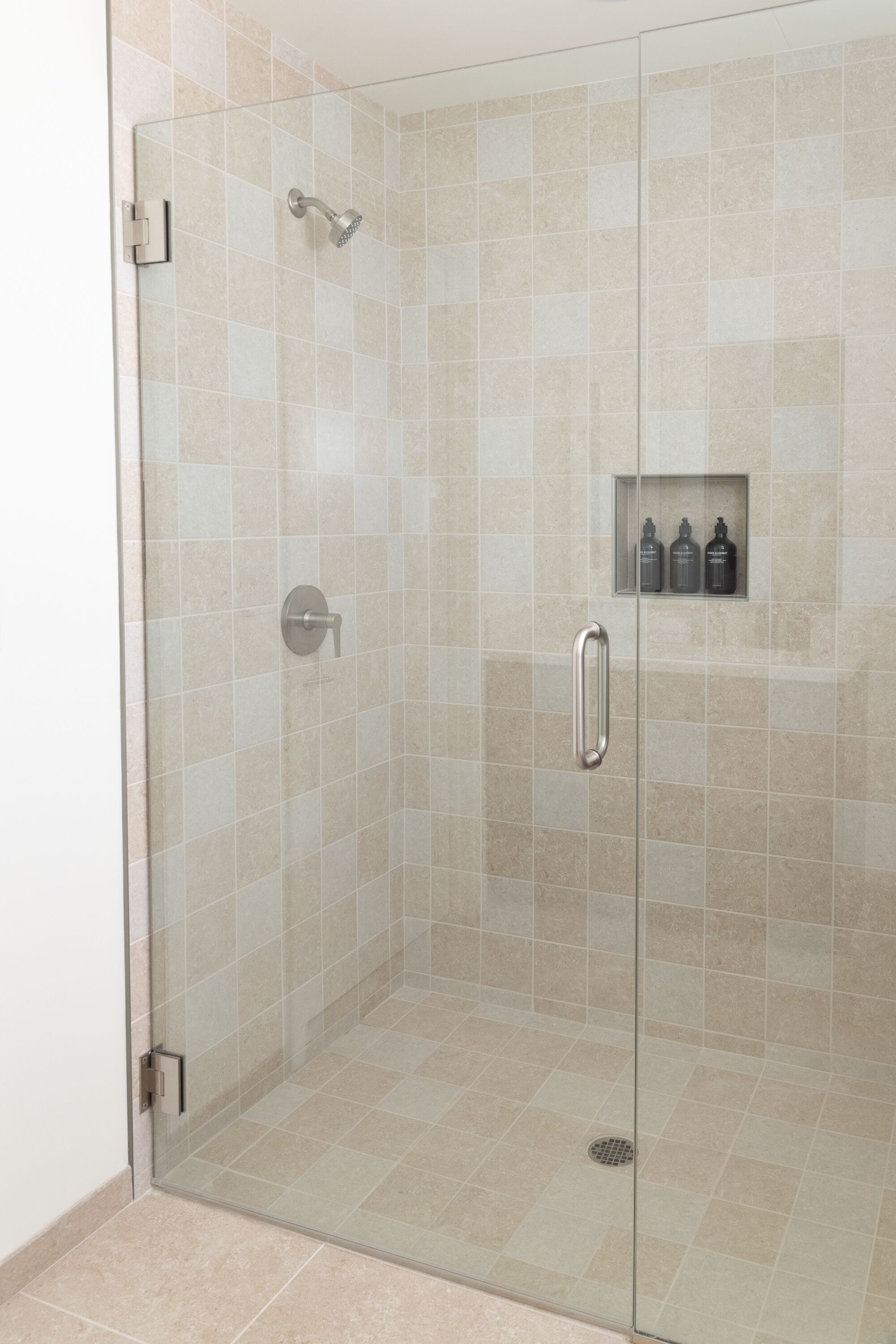 A glass-enclosed shower area with beige tiles, a handheld showerhead, and a shelf with toiletries.