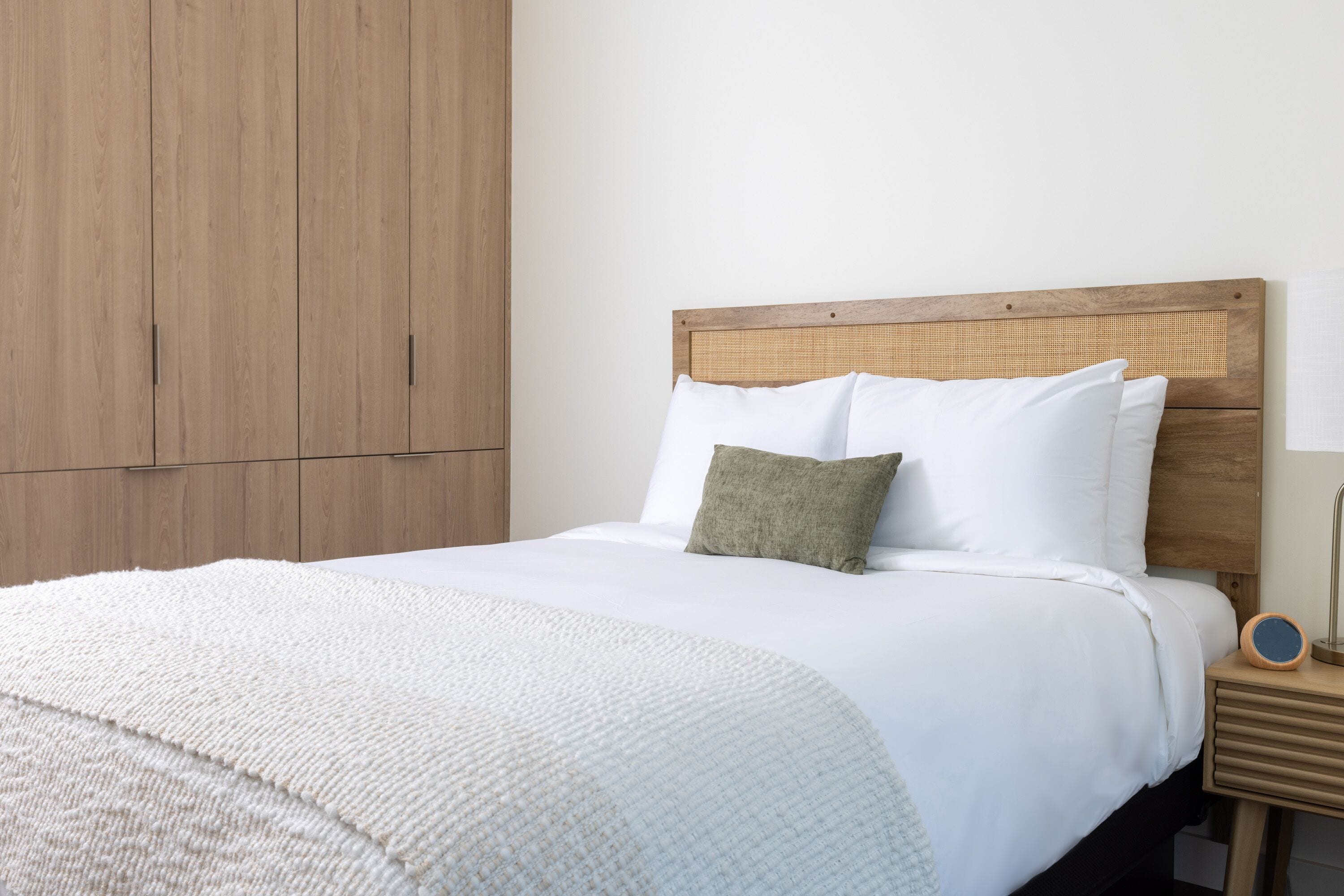 Una cama bien hecha con ropa blanca y un almohadón verde, un cabecero de madera y una mesita de noche con una lámpara.