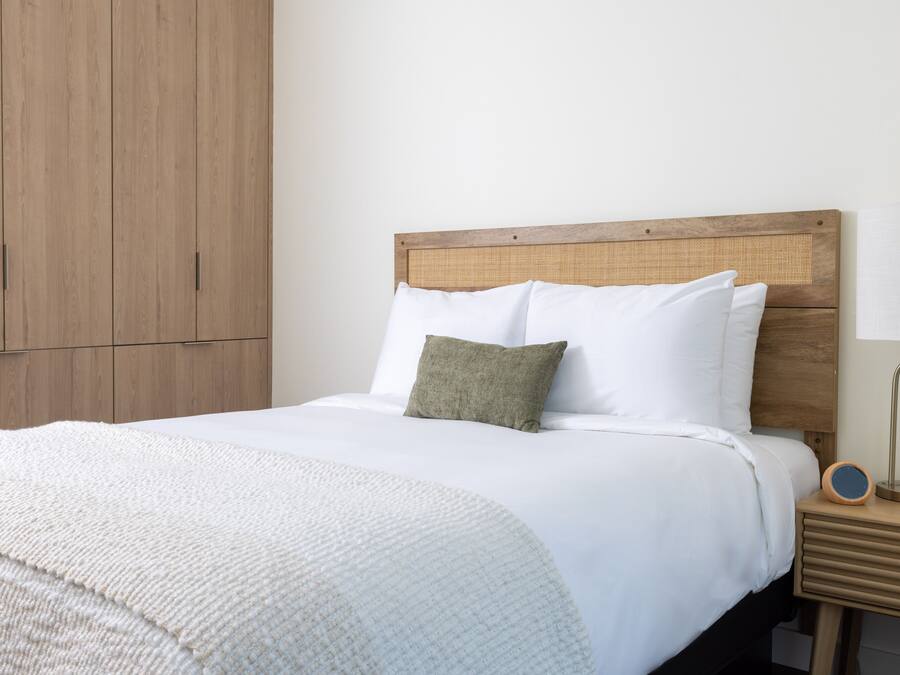 Una cama bien hecha con ropa blanca y un almohadón verde, un cabecero de madera y una mesita de noche con una lámpara.