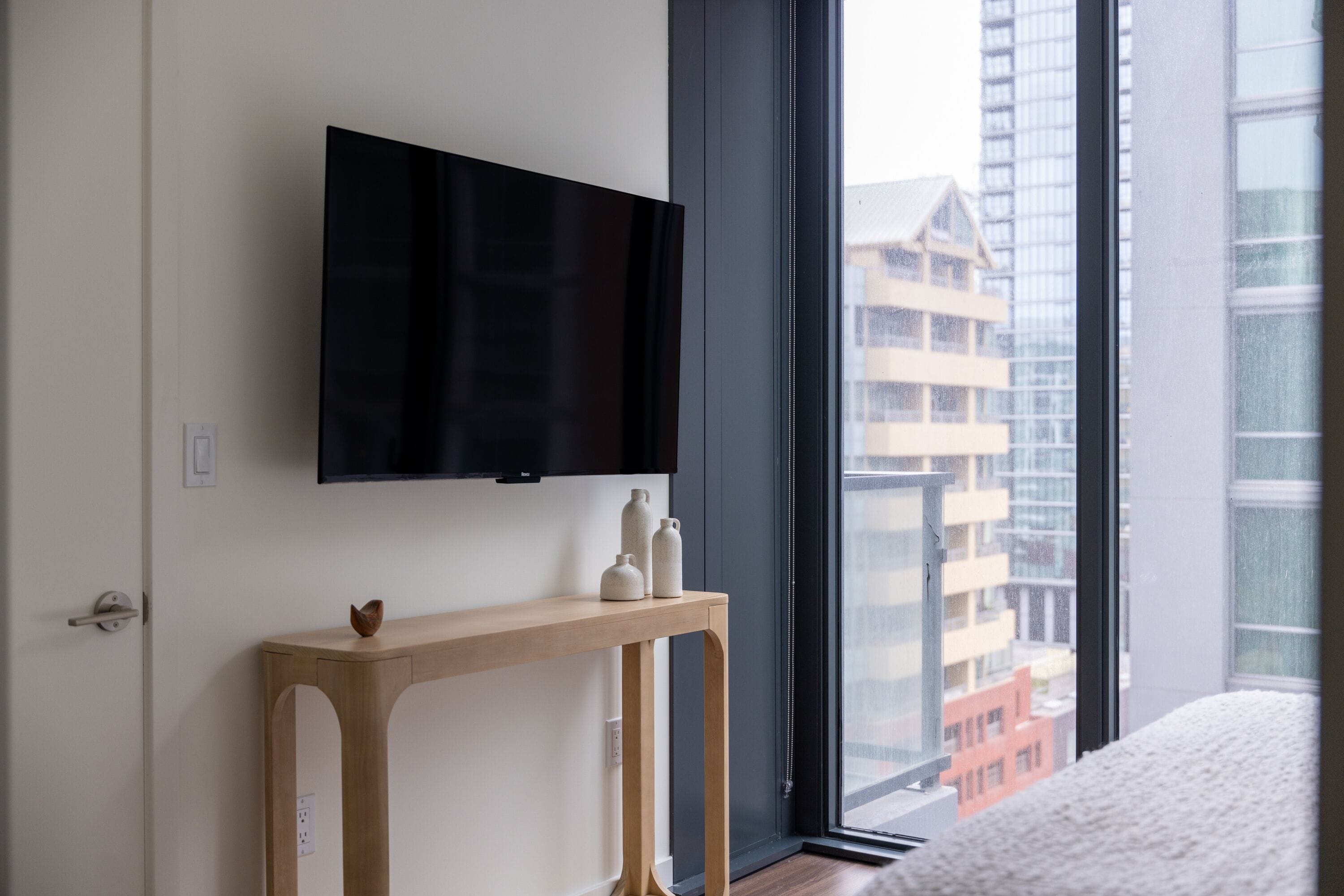 A modern hotel room with a large window, a flat-screen TV mounted on the wall, and a wooden console table with decorative items.