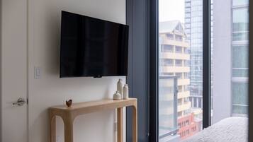 A modern hotel room with a large window, a flat-screen TV mounted on the wall, and a wooden console table with decorative items.