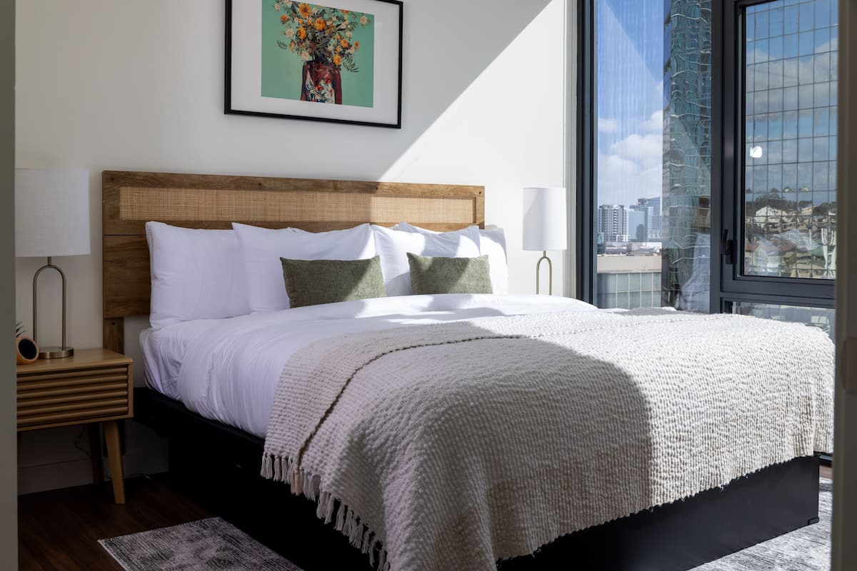 Een moderne slaapkamer met een groot bed, een houten hoofdbord, een kunstwerk in een lijst erboven en een stadszicht door een groot raam.