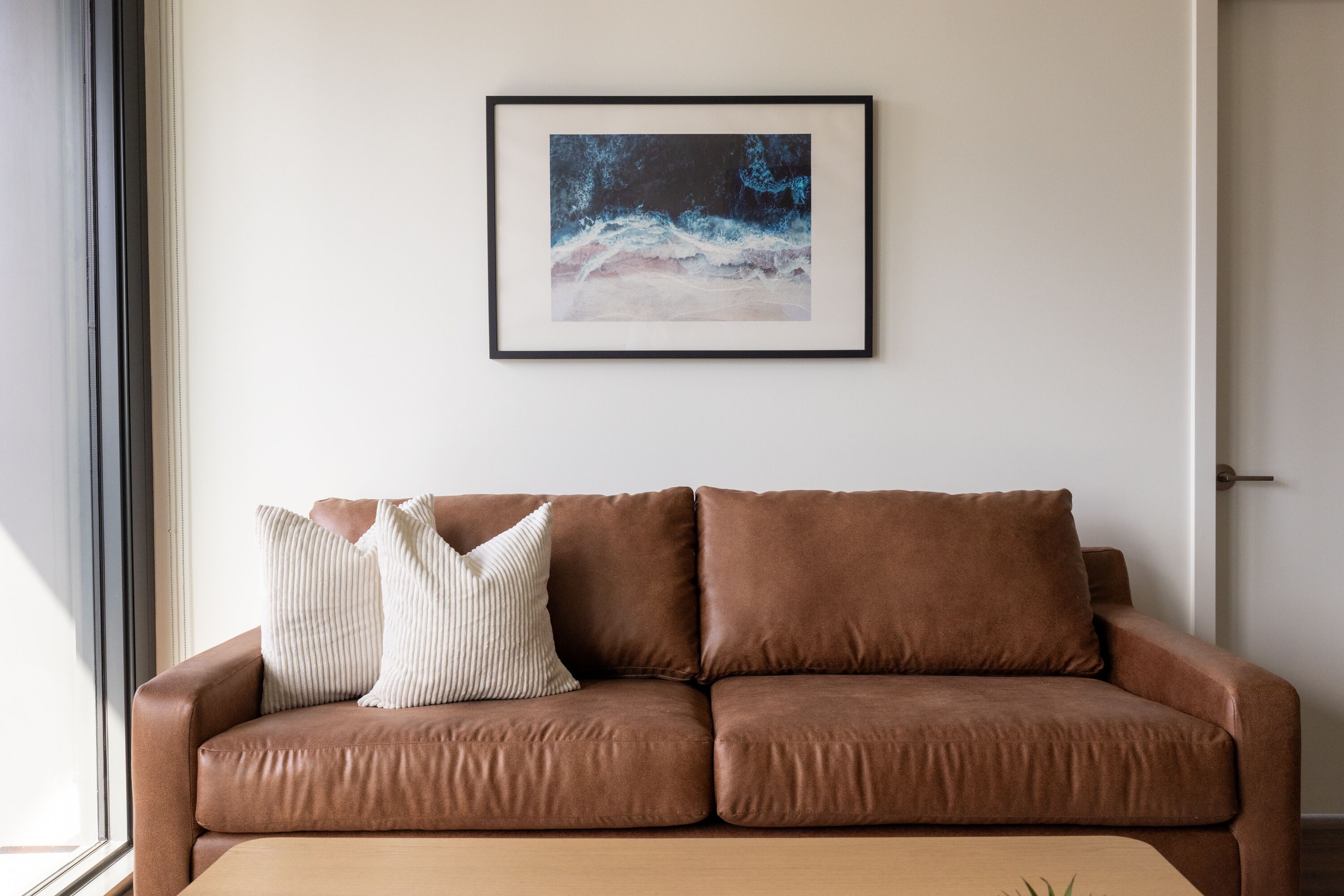 A brown leather sofa with two pillows against a white wall, a framed picture above it, and a window to the left.