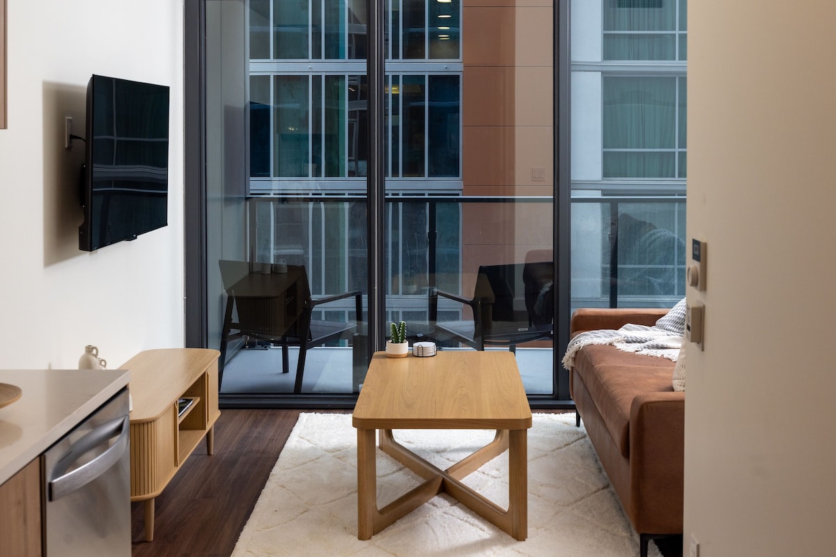 Una sala de estar moderna con una mesa de centro de madera, un sofá, un televisor montado en la pared y un ventanal con vistas a la ciudad.