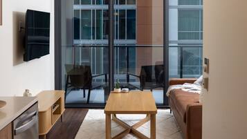 A modern living room with a wooden coffee table, a sofa, a TV mounted on the wall, and a large window offering a view of the city.