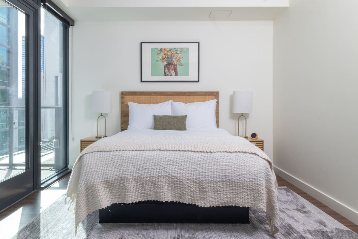 Een moderne slaapkamer met een groot bed, twee nachtkastjes, een ingelijst kunstwerk boven het bed, en uitzicht door een groot raam.