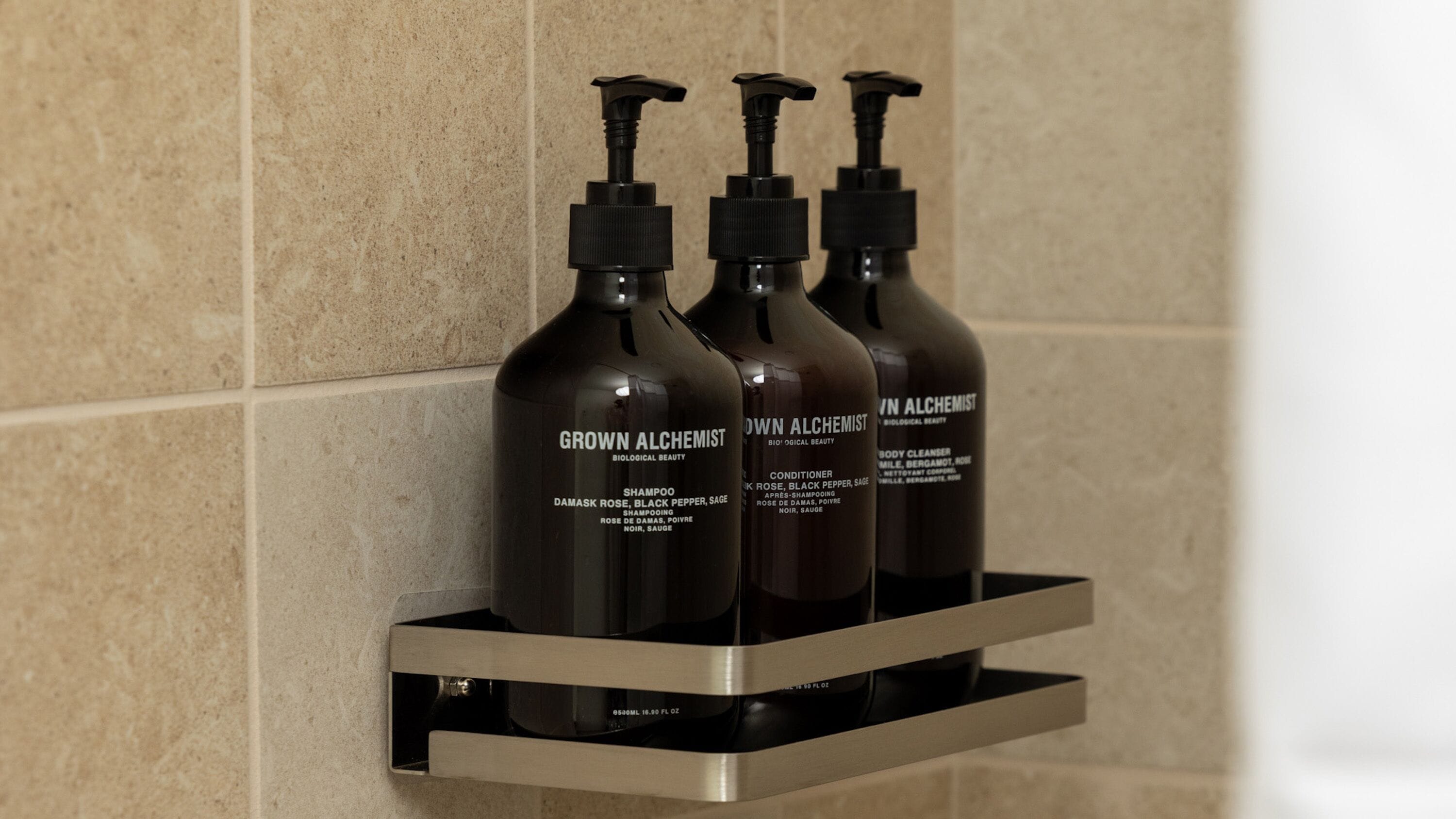 Three bottles of 'Grown Alchemist' products on a metal shelf against a tiled wall.