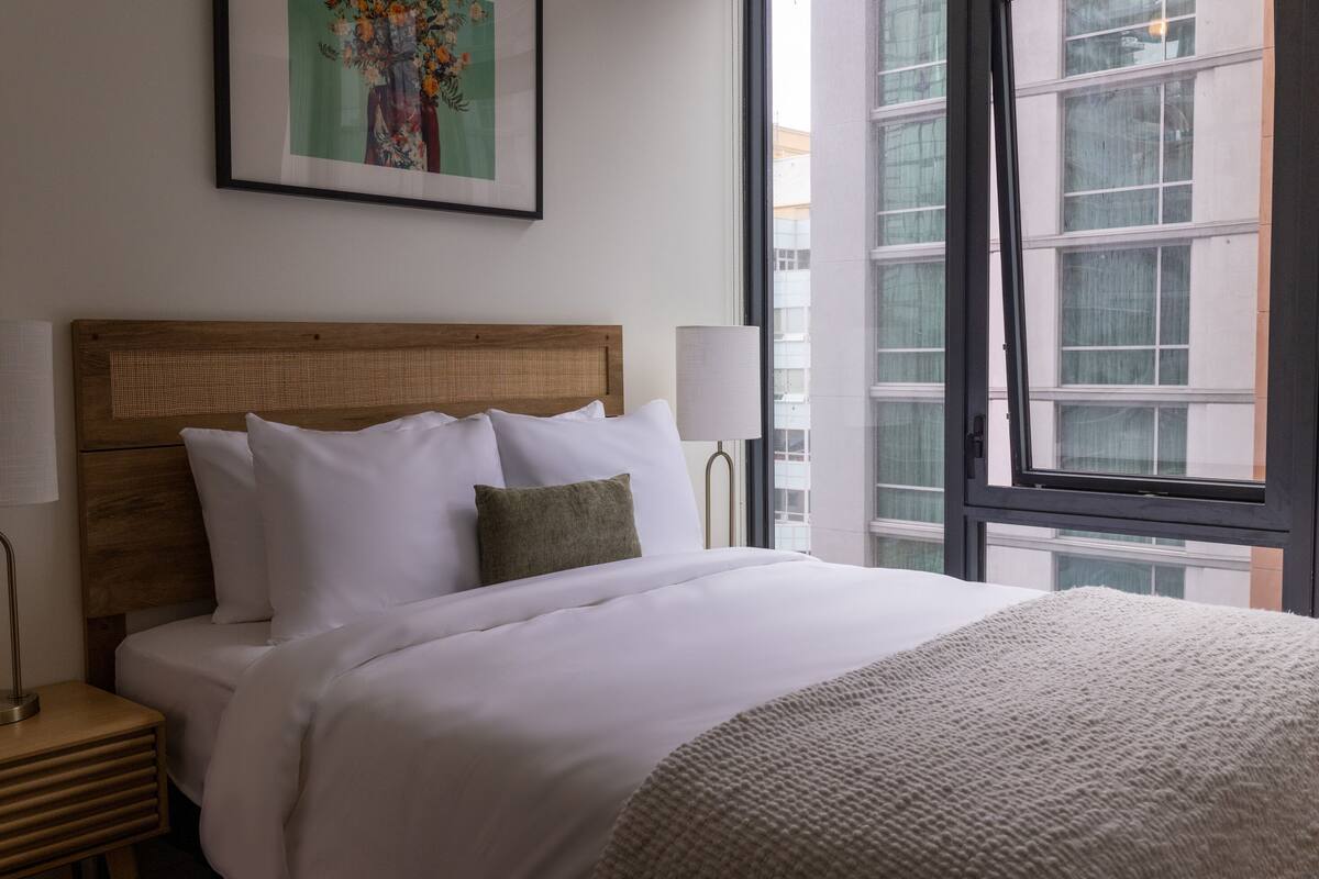 Een net opgemaakt bed met witte lakens en een gebreid plaid, een houten hoofdbord, een bedlampje en een lijst met bloemmotieven aan de muur.