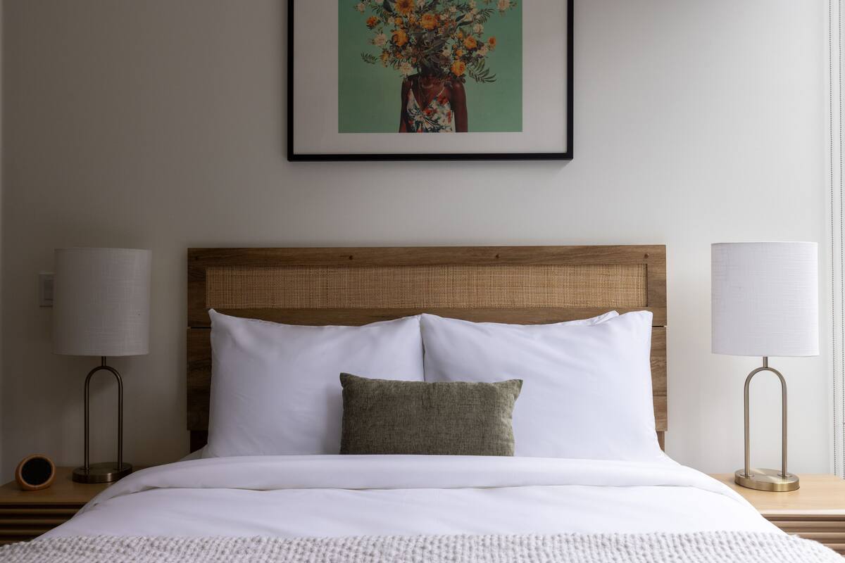 Een opgemaakt bed met witte lakens, een houten hoofdbord en een enkel groen kussen. Er staan twee bedlampen op houten nachtkastjes.